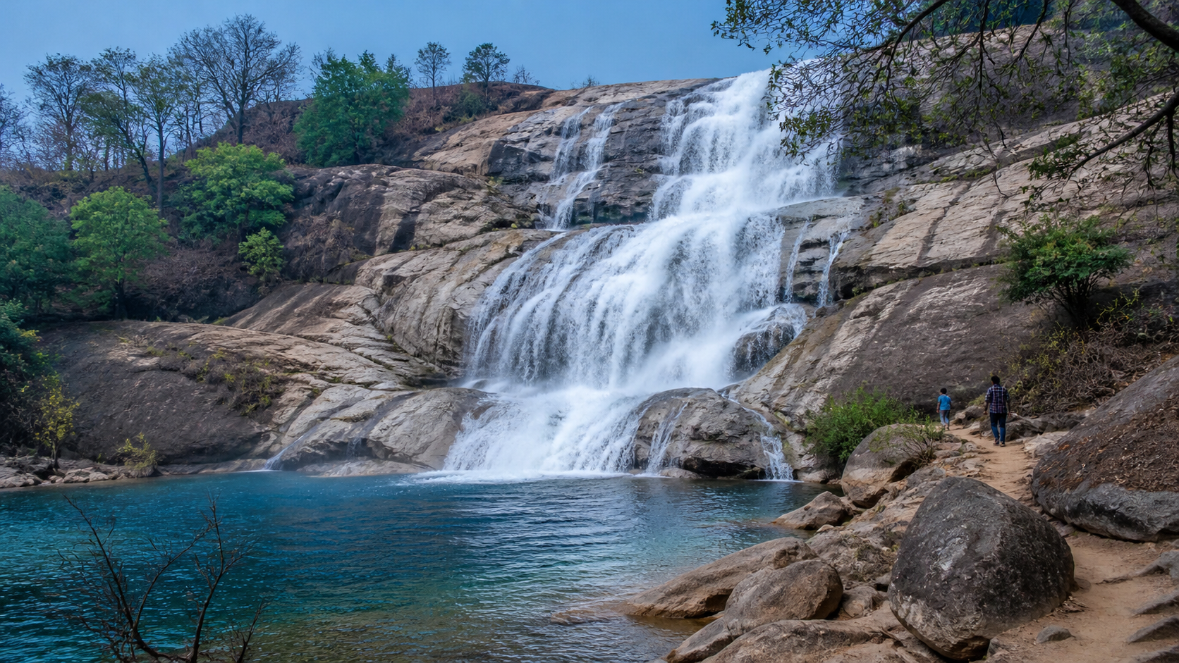 A bright waterfall flowing over pale rock into a blue pool in rural India